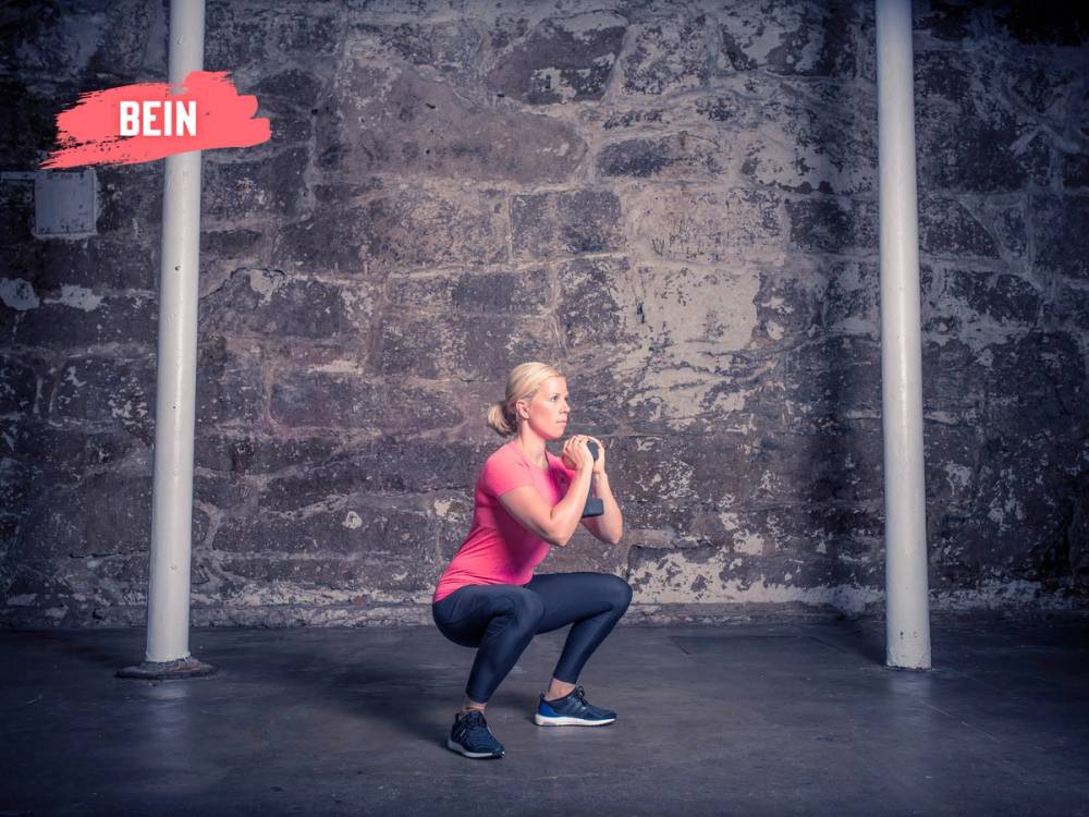 En kvinne gjør en knebøyøvelse foran en murvegg, iført rosa treningstøy og svarte sko.