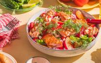 En stor bolle med en fargerik salat som inneholder tomater, agurk, urter og sitronbåter, omgitt av drikker og andre matvarer på et bord.