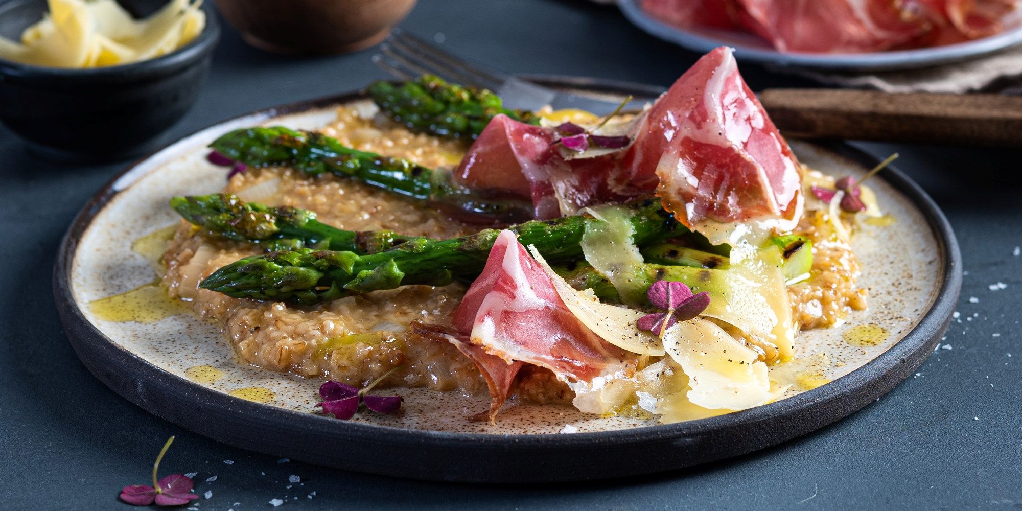 Havreotto med asparges og coppa servert på en tallerken med grønne urter og blomsterdekorasjon, sett fra siden.