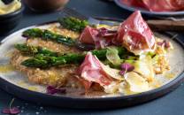 Havreotto med asparges og coppa servert på en tallerken med grønne urter og blomsterdekorasjon, sett fra siden.