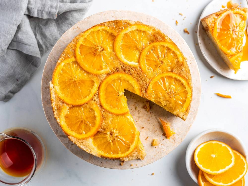 En opp ned appelsinkake med skiver av appelsin på toppen, en skive tatt ut av kaken, og små biter av kaken rundt på en hvit overflate.