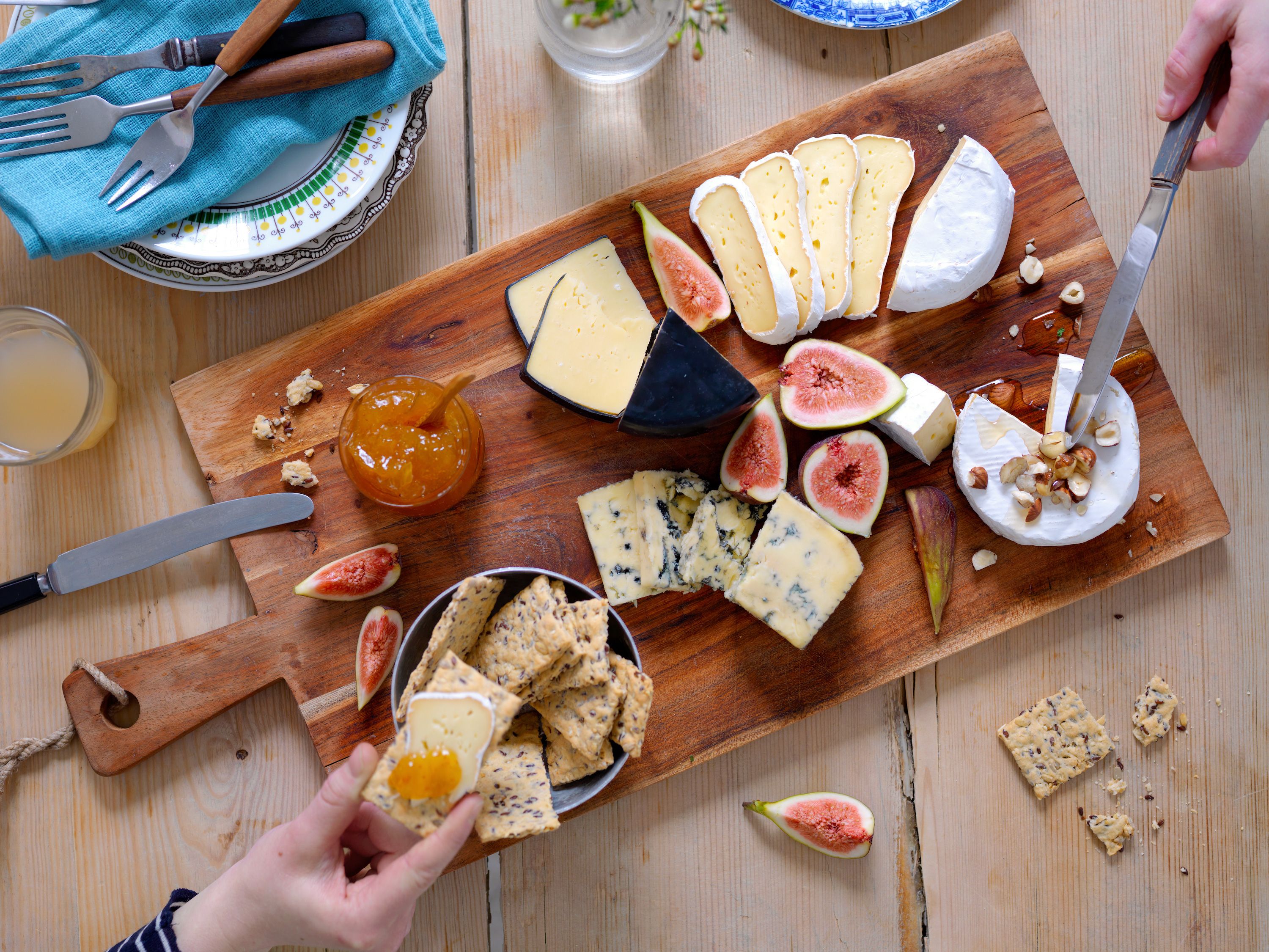 Et ostefat med ulike typer ost, fiken og kjeks på et trebord, med en person som skjærer ost og en annen som tar en bit av en kjeks.