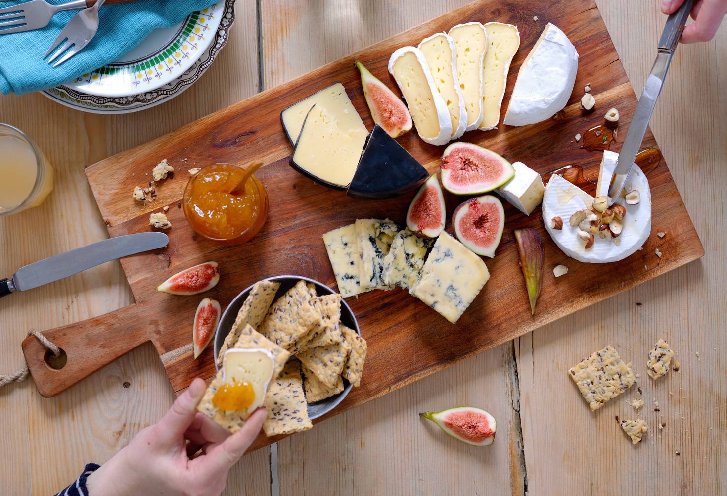 Et ostefat med ulike typer ost, fiken og kjeks på et trebord, med en person som skjærer ost og en annen som tar en bit av en kjeks.