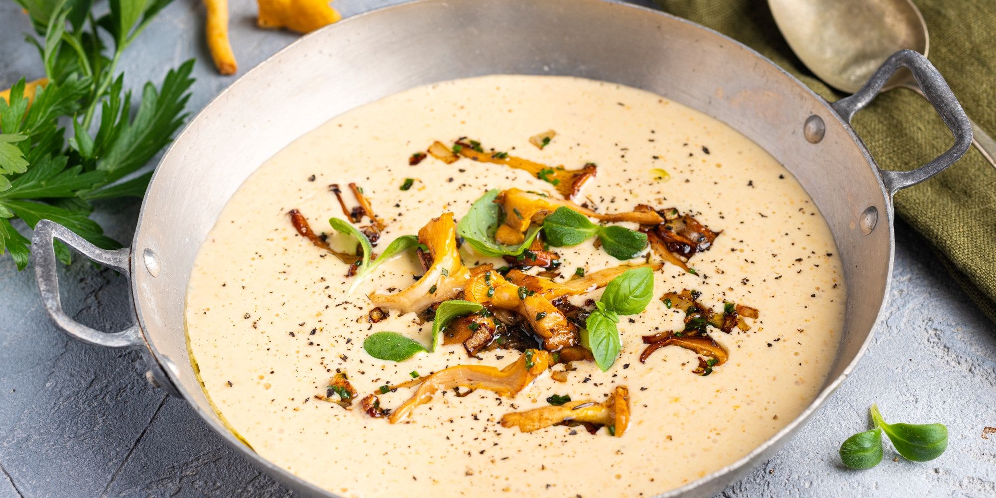 Kremet soppsuppe servert i en bolle med soppbiter og urter på toppen, omgitt av grønne urter og en skje.