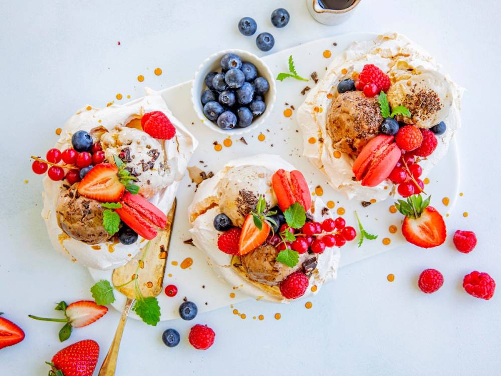 Tre porsjoner med pavlova toppet med bær og frukt, og en skål med blåbær i midten