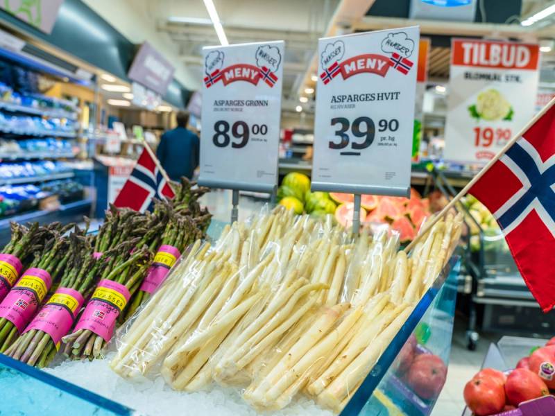 Asparges hvit og grønn på en matbutikk, med norske flagg og prismermer i bakgrunnen.