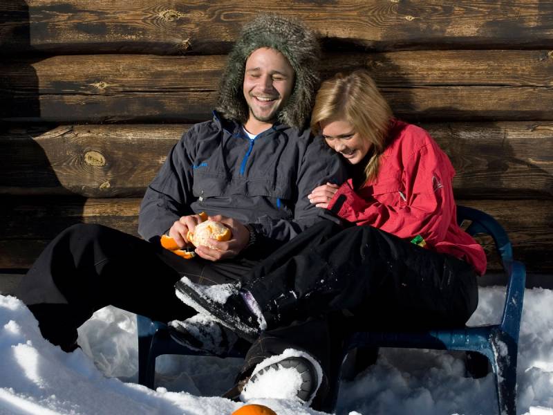 To personer sitter på en benk i snøen, med en bakgrunn av trevegger, og de har på seg vinterklær, mens de spiser mat.