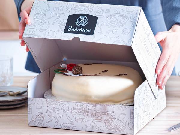 Person åpner en hvit bakeboks med en ferdiglaget kake inni på et bord.