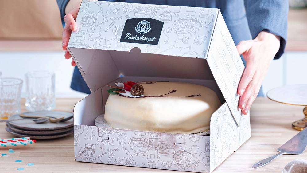 Person åpner en hvit bakeboks med en ferdiglaget kake inni på et bord.