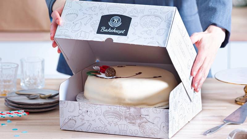 Person åpner en hvit bakeboks med en ferdiglaget kake inni på et bord.