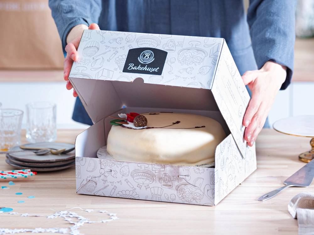 Person åpner en hvit bakeboks med en ferdiglaget kake inni på et bord.