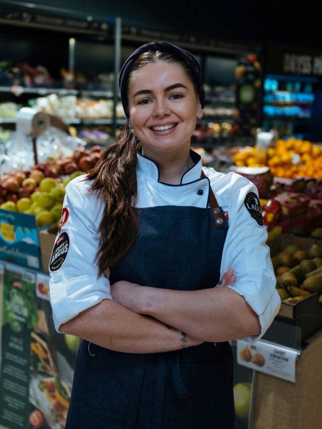 En person i kokkeforkle med lang, mørk hår står i en matbutikk eller frukt- og grøntavdeling med frukt og grønnsaker i bakgrunnen.