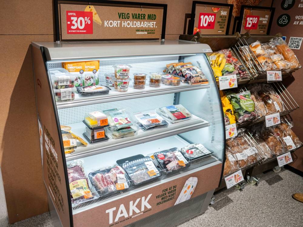 En kjøledisk med ulike pakkede kjøtt- og påleggprodukter i en dagligvarebutikk, med reklameskilt i bakgrunnen.