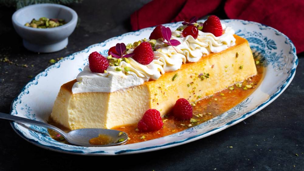 Karamellpudding med krem og friske bringebær på en oval tallerken med blå mønster.