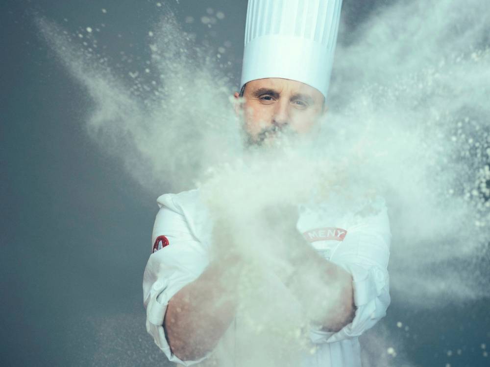 En person kaster mel opp i luften, og melbølger sprer seg rundt dem, med en tydelig melhåndtering i fokus.