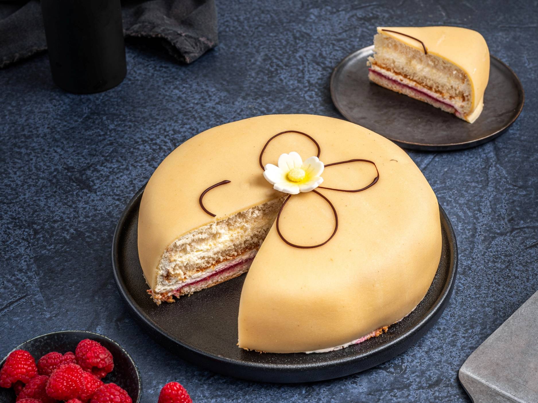 En rund kake dekket med gul fondant og pyntet med en enkel blomst og kurver av sjokolade, med et stykke tatt ut på en tallerken i bakgrunnen.