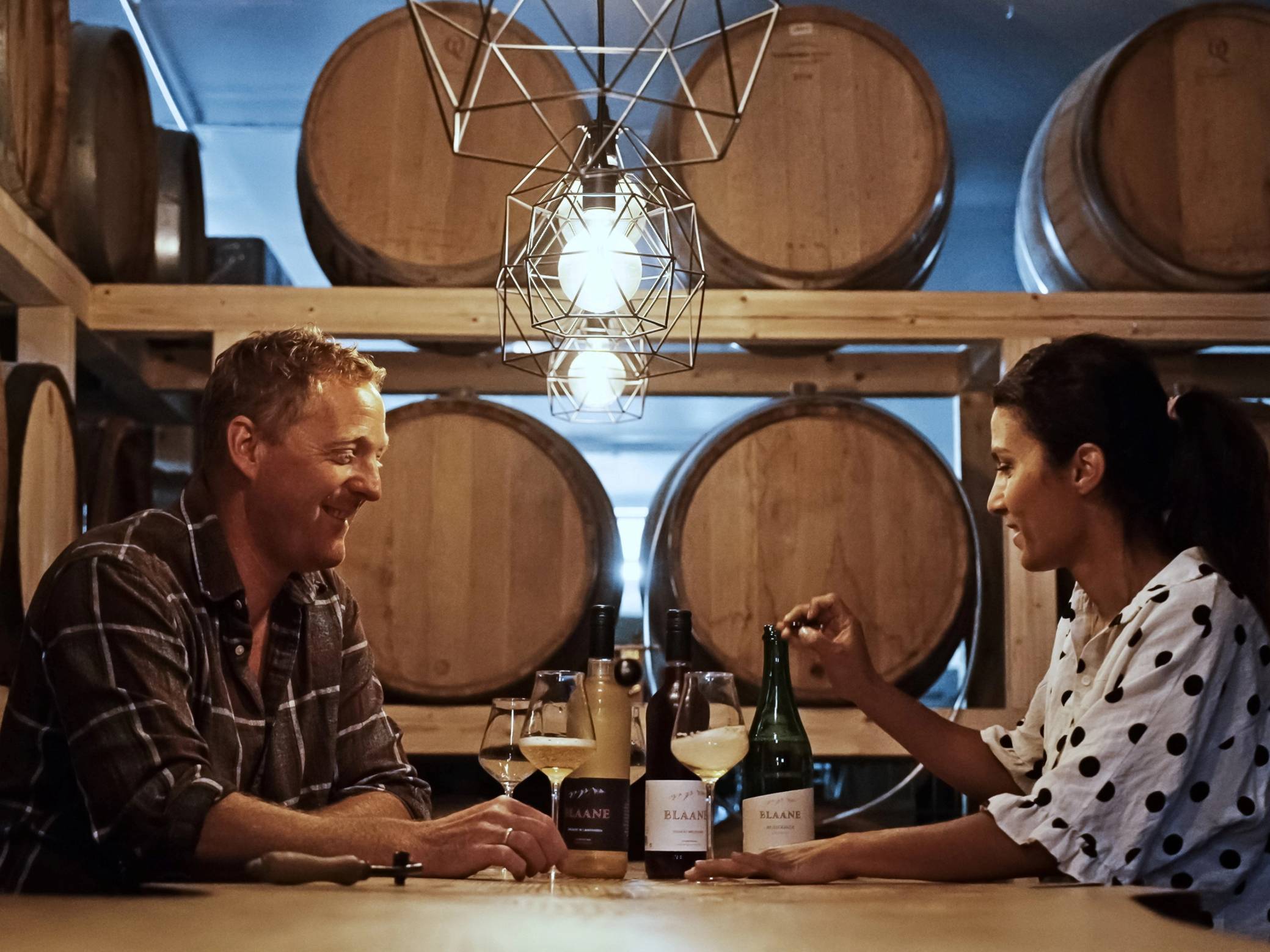 To personer som sitter ved et bord i en vinkjeller, med store vinfat på veggen bak og vinflasker foran dem, mens de snakker og har et glass vin hver.