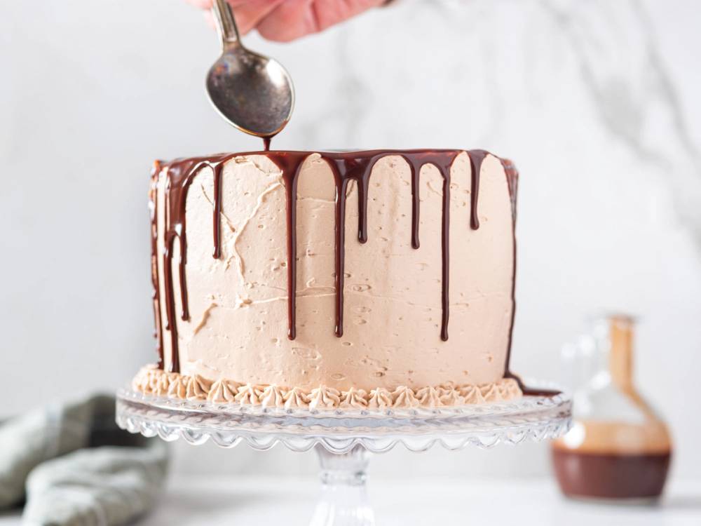 En kake med hvit krem og sjokoladedrips som drypper nedover sidene, plassert på et glasskakefat.