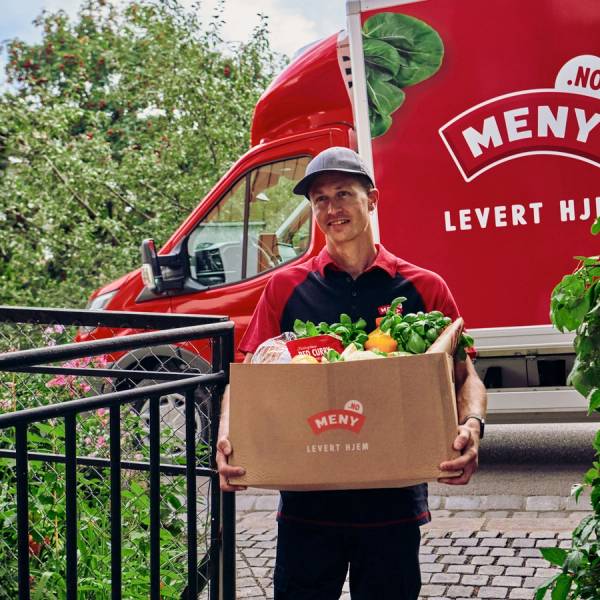 En person med ansiktet dekket til, som bærer en eske med matvarer foran en rød matbil med logo og teksten 'Levert Hjem', omgitt av grønne busker.