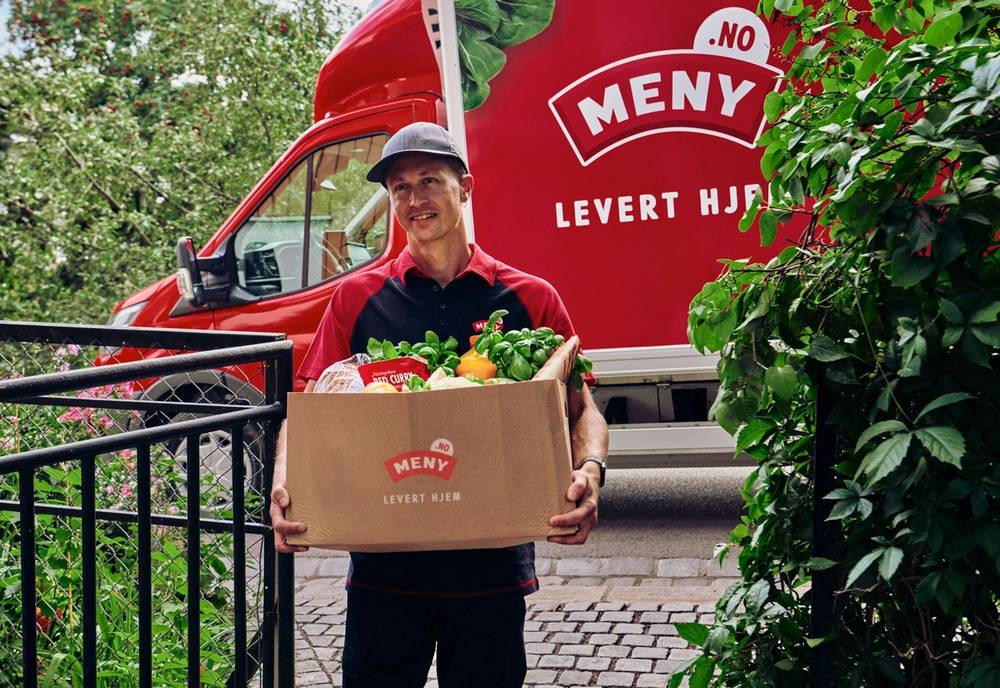 En person med ansiktet dekket til, som bærer en eske med matvarer foran en rød matbil med logo og teksten 'Levert Hjem', omgitt av grønne busker.
