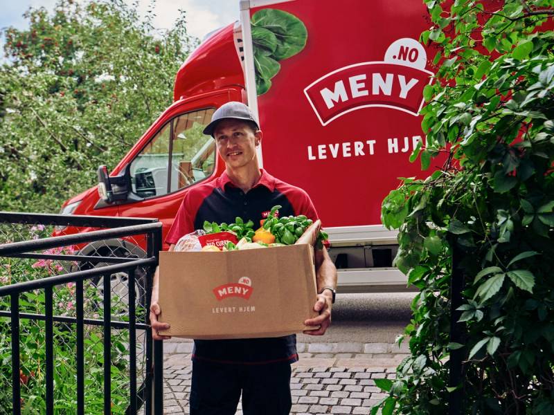 En person med ansiktet dekket til, som bærer en eske med matvarer foran en rød matbil med logo og teksten 'Levert Hjem', omgitt av grønne busker.