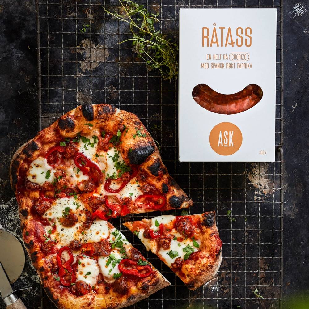 En pizza med chorizo, paprika og ost på en svart rist, med en eske med chorizo ved siden av.