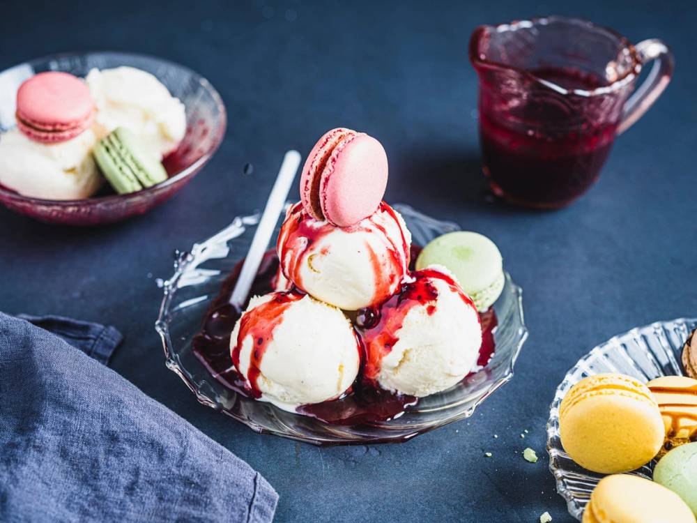 Iskuler med ulike smaker og farger, servert i en glassbolle med en skje, og en kopp med rød saft i bakgrunnen.