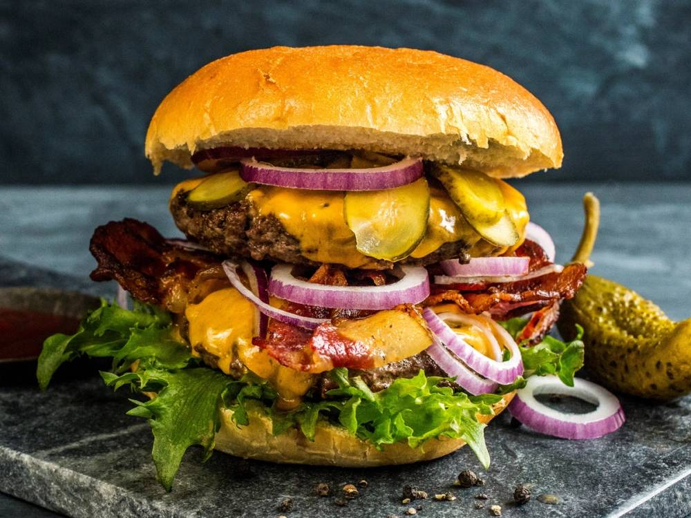 En saftig burger med kjøttkake, ost, bacon, løk, sylteagurk og salat i et brød, med en liten skive sylteagurk ved siden av.