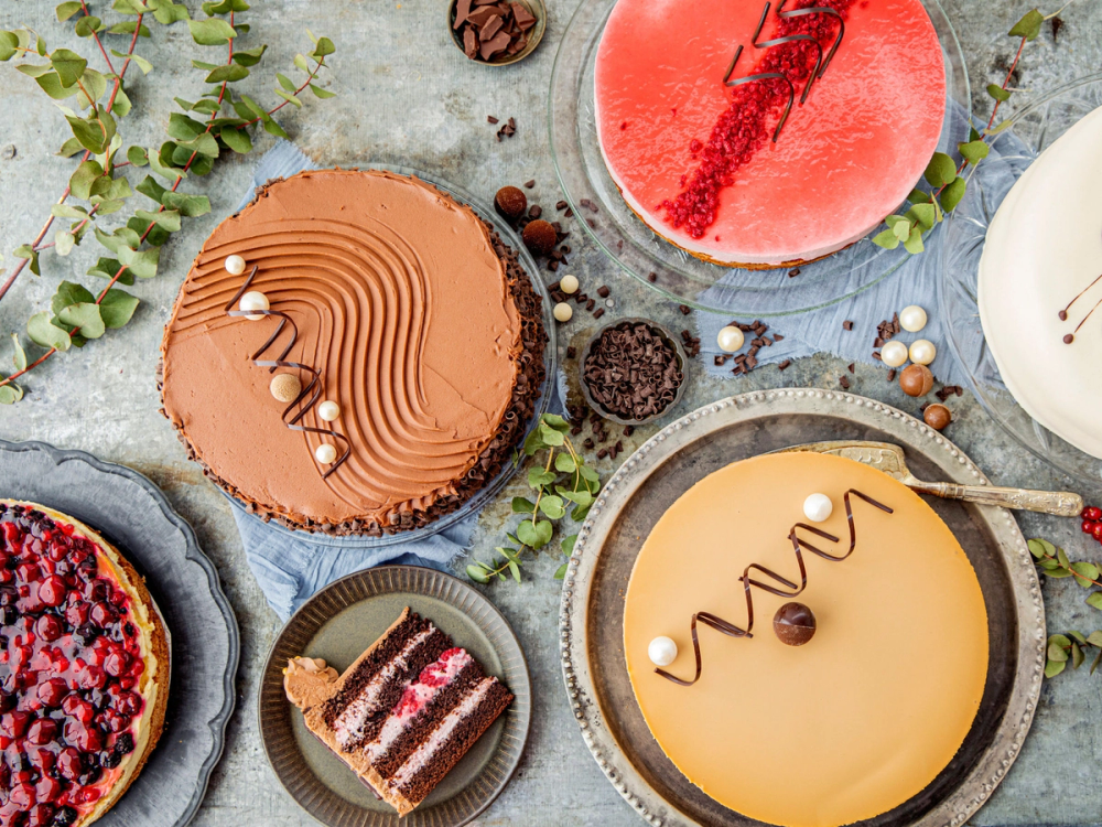 Ulike kaker og desserter på et bord, inkludert en sjokoladekake, en sitronkake og en vannmelon, omgitt av grønne blader og små dekorasjoner.