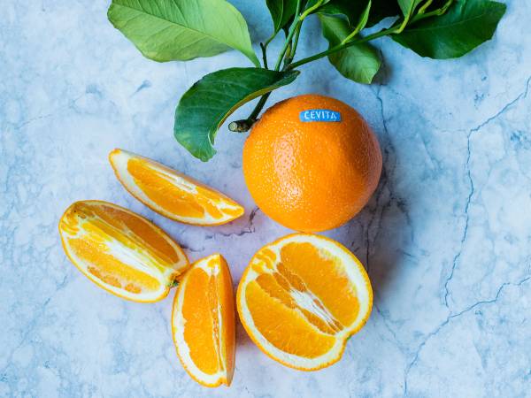 En hel appelsin med grønne blader og flere oppkuttede appelsinbiter på en lys blå bakgrunn.