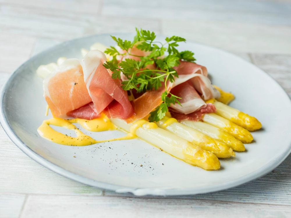 Hvit asparges med sitronhollandaise og skinke på en tallerken, pyntet med frisk grønn urter.
