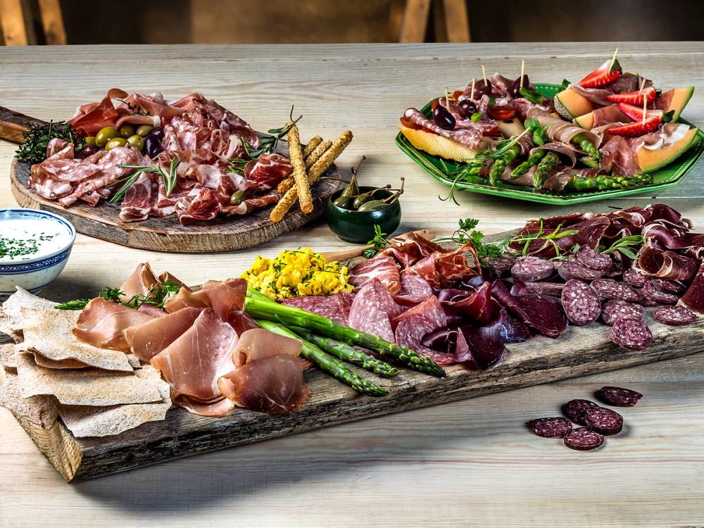 Et spekefat med ulike typer kjøttpålegg, grønnsaker og brødskiver på et treplate, klar til servering.