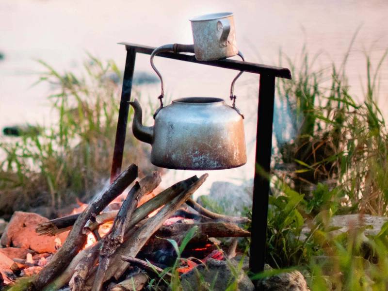 En gammel kjele henger over et bål på en strand, med en kopp plassert på en enkel ramme over kjelen.
