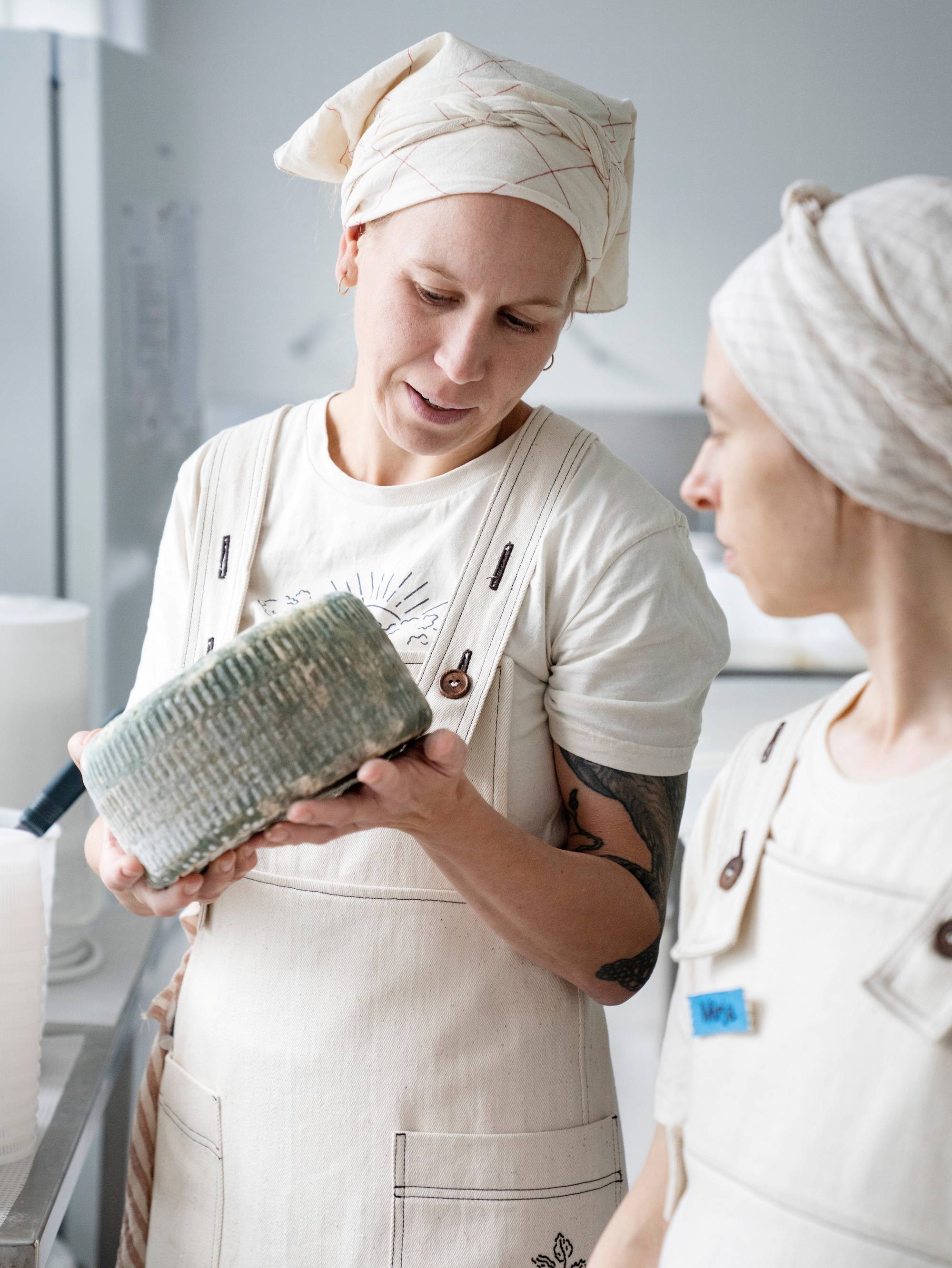 To personer iført kokkeuniformer og hodeplagg står i et kjøkken, hvor den ene holder en stor osteklokke.