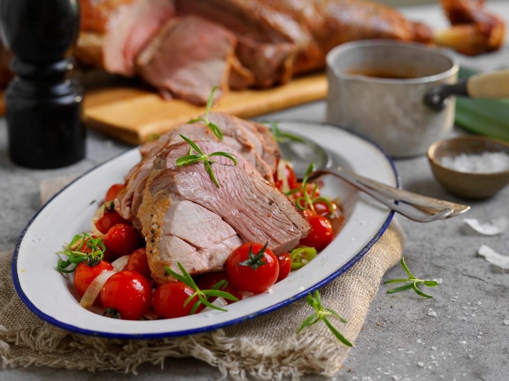 Lammelår med cherrytomater og tzatziki på en hvit tallerken, omgitt av urter og kjøkkenutstyr.