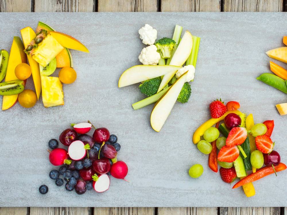 Ulike frukt- og grønnsaksbiter arrangert på en lys treplate, inkludert druer, jordbær, agurk, gulrot og eple