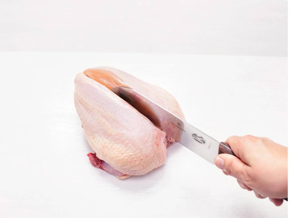 En person skjærer i en hel rå kylling med en kniv på et hvitt bord.