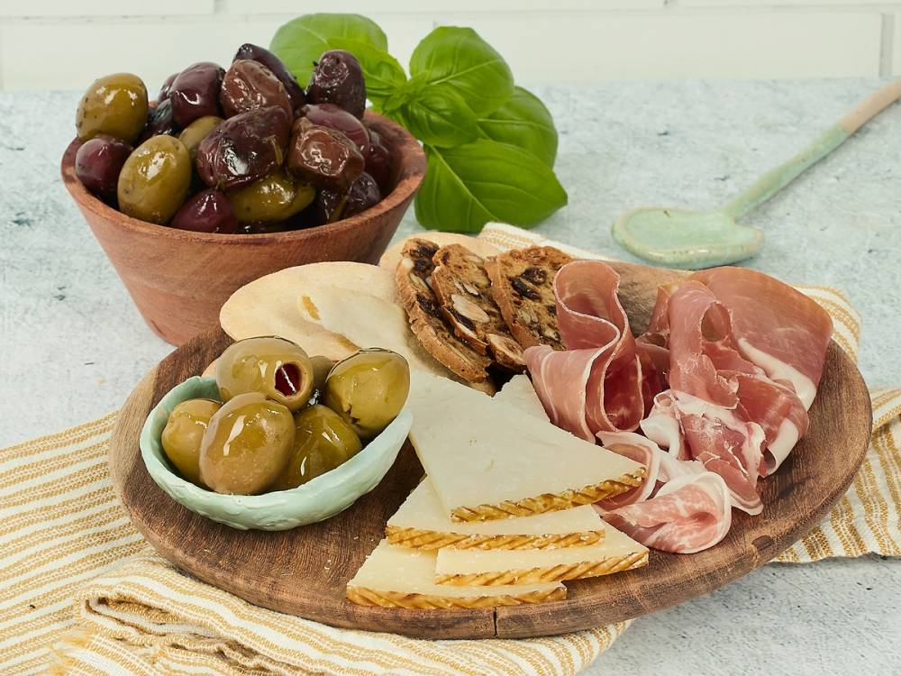 Et fat med ulike typer oliven, ost og skinke på et trefat, med en grønn basilikumplante i bakgrunnen.