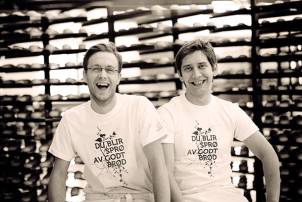 To personer står i en butikk eller bakeri med hyller fylt med brød i bakgrunnen, begge iført hvite t-skjorter med tekst.