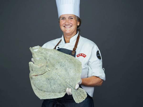 En person i kokkeforkledning holder en stor, flat fisk foran kroppen, med ansiktet utelatt.