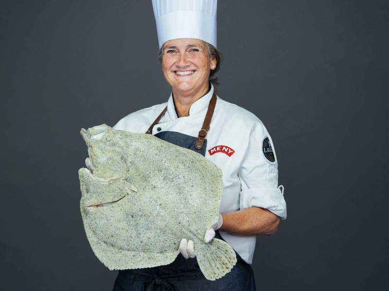 En person i kokkeforkledning holder en stor, flat fisk foran kroppen, med ansiktet utelatt.
