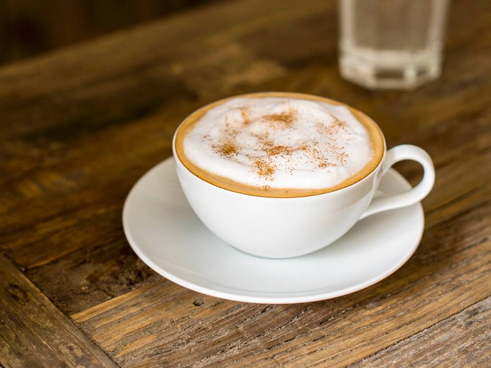 En kopp cappuccino med skum og kanel på toppen på et treverkbord, med et glass vann i bakgrunnen.