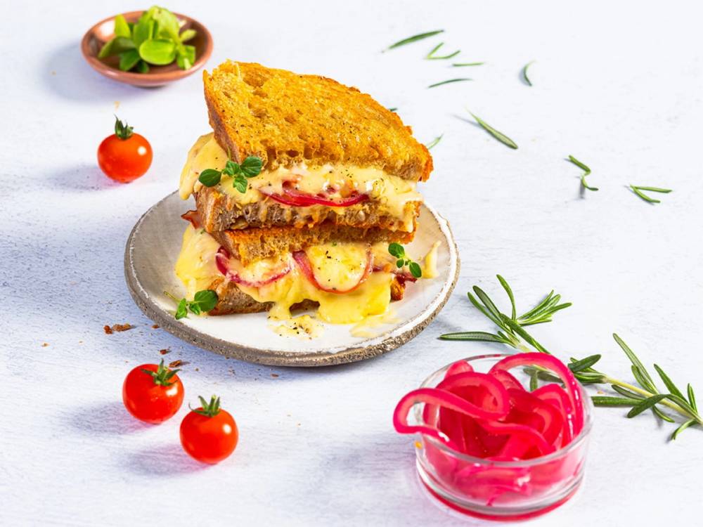 To toast med ost og skinke på en tallerken, omgitt av cherrytomater og urter, med en liten skål med rosa straws i bakgrunnen.