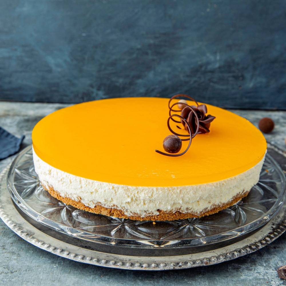 En ostekake med en glatt, gul overflate pyntet med sjokoladebiter og sjokoladespiraler på en sølvfat.
