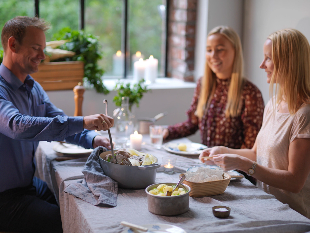 Tre personer spiser fårikål rundt et spisebord i et lyst rom med store vinduer og grønne planter.