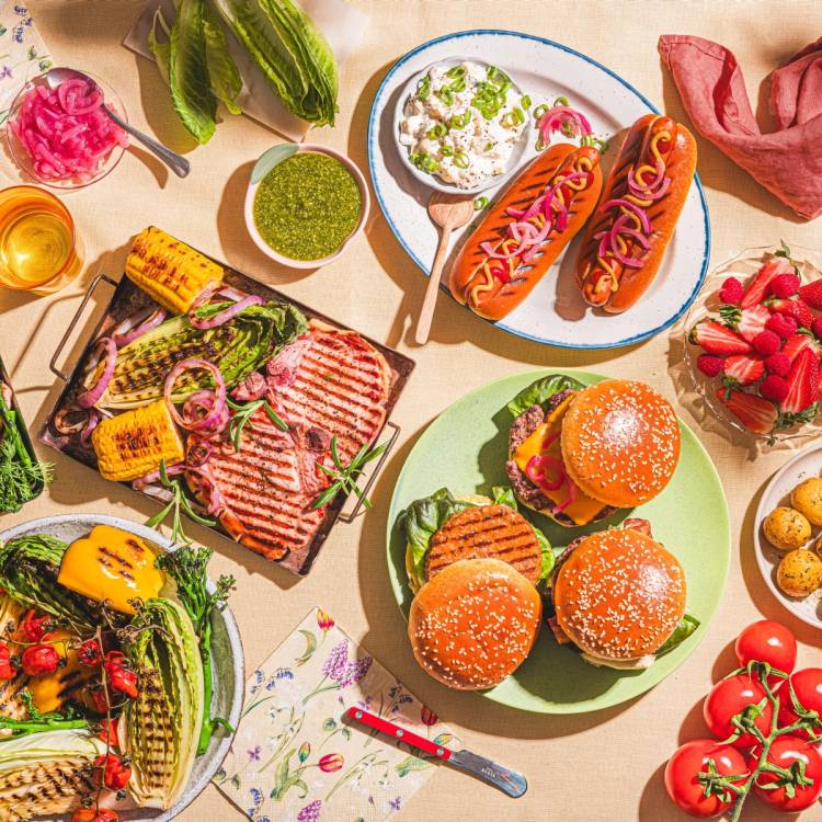 Et utvalg av mat og snacks, inkludert burgere, pølser, kaker og grønnsaker, er arrangert på et bord til en sommerfest.