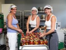 Tre kvinner i et kjøkken lager syltetøy med frukt og grønnsaker, alle iført kokkeuniformer og hårnetter.