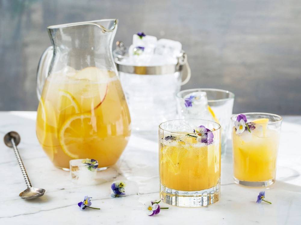 En glass og en mugge med eplejuice-basert mocktail punch med is og blomsterdekorasjoner på et lyst bord.