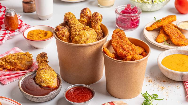 Friterte kyllingbiter i papirbeger omgitt av ulike sauser og salater på et bord.