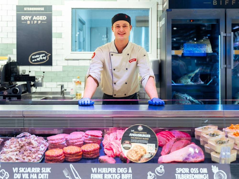 En kok står bak en kjøttdisk med ulike typer kjøtt og pølser i en butikk, med en fryser til høyre og reklameskilt foran.